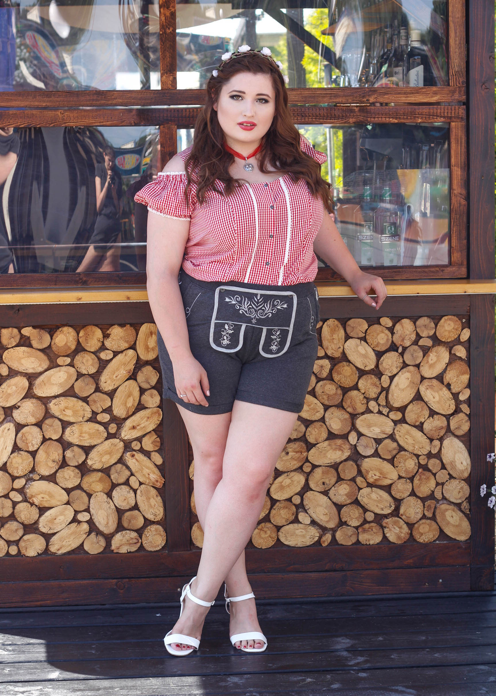 Junge kurvige Frau vor einer Holzhütte im Oktoberfest Outfit. Plus Size Lederhosen Look.